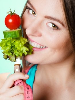 Eating, dieting, slimming and healthcare concept. Young fit woman with measuring tape on neck holding and biting fresh mixed vegetables. Studio shot on blue background.