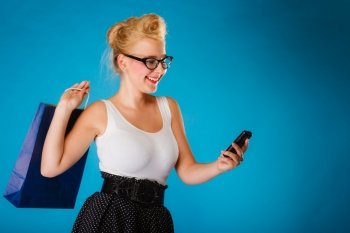 Retro style. Young woman pinup girl in glasses with shopping bag using on cell phone. Blue background.