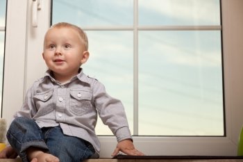 Happy childhood. Portrait of smiling little boy at home. Cute child kid in blue shirt sitting on the windowsill. Copy space.