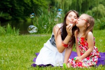 Family happiness and carefree concept. Mother and daughter little girl having fun blowing soap bubbles together in park, green blurred background
