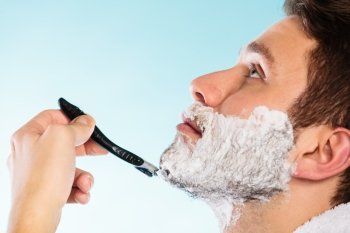 Health beauty and skin care concept. Closeup young bearded man with foam on face shaving with razor on blue background.