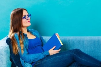 Leisure, education, literature and home concept - Woman in glasses wearing blue denim sitting on couch reading book at home