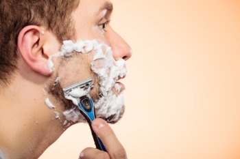 Health beauty and skin care concept. Closeup young bearded man with foam on face shaving on bright orange background.