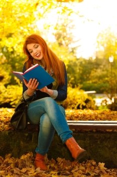 Fall lifestyle concept. Redhaired young woman girl relaxing in autumn park reading book, sitting alone on bench. Sunny day